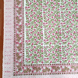 May include: A white textile with a repeating floral pattern in pink and green. The border features a geometric design in pink. The overall design is a floral print, suitable for home decor.