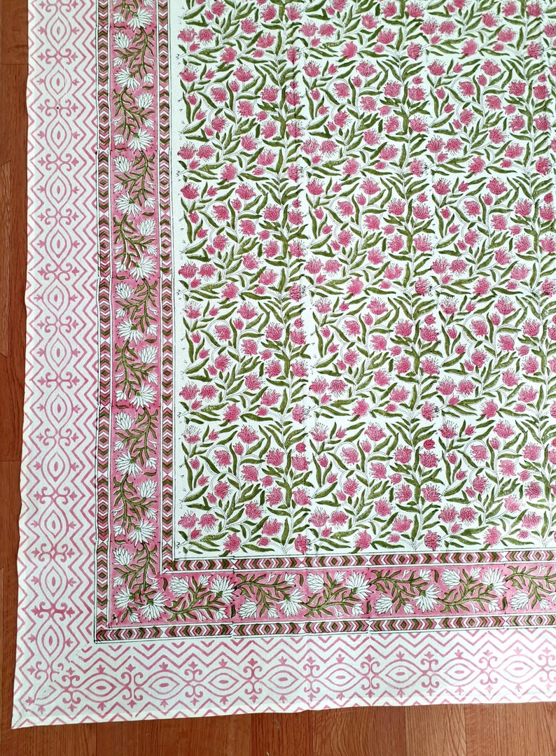 May include: A white textile with a repeating floral pattern in pink and green. The border features a geometric design in pink. The overall design is a floral print, suitable for home decor.