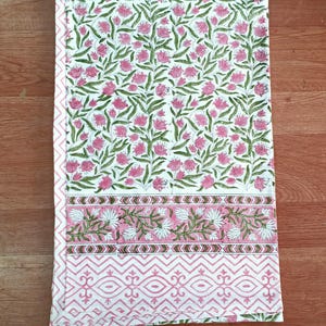 May include: A white tablecloth with a floral pattern of pink flowers and green leaves. The border features a pink and white geometric design. The tablecloth is folded and lies on a wooden surface.
