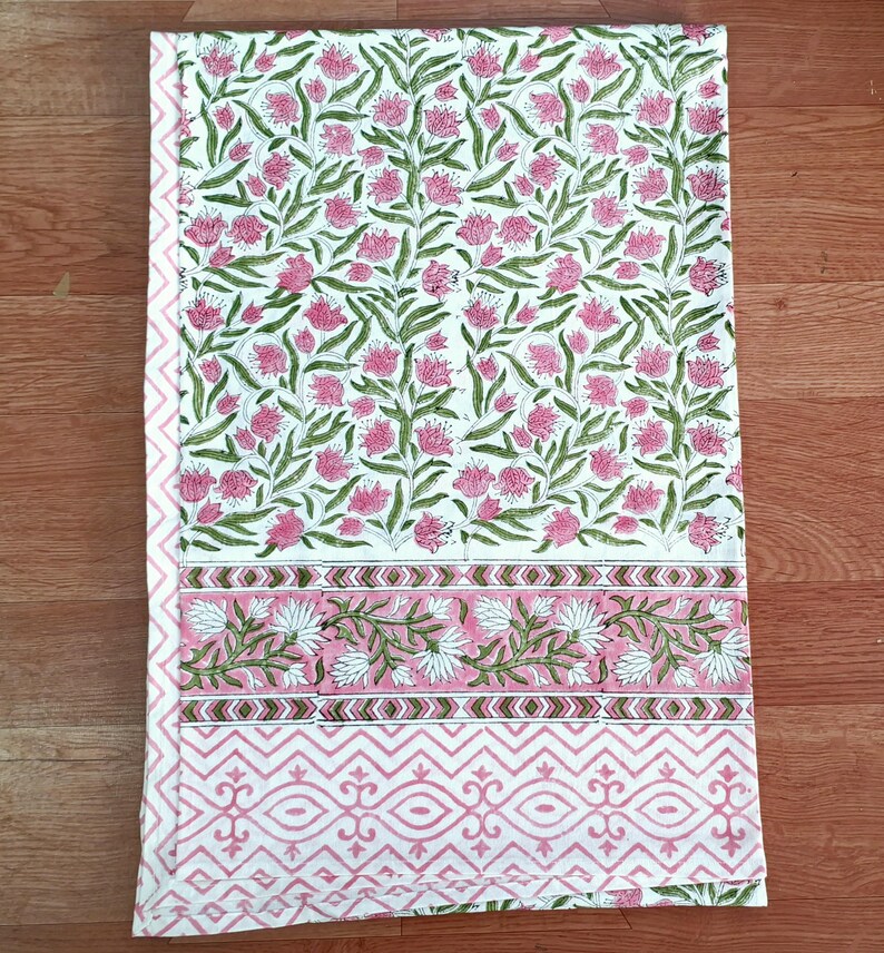 May include: A white tablecloth with a floral pattern of pink flowers and green leaves. The border features a pink and white geometric design. The tablecloth is folded and lies on a wooden surface.