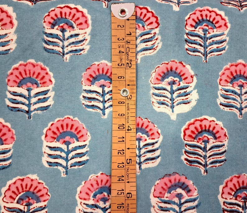 May include: Pale blue fabric featuring a repeating floral design in pink, white, and blue. A wooden ruler with black markings and the text "MADE IN INDIA" is placed on the fabric. The ruler measures up to 15 cm.