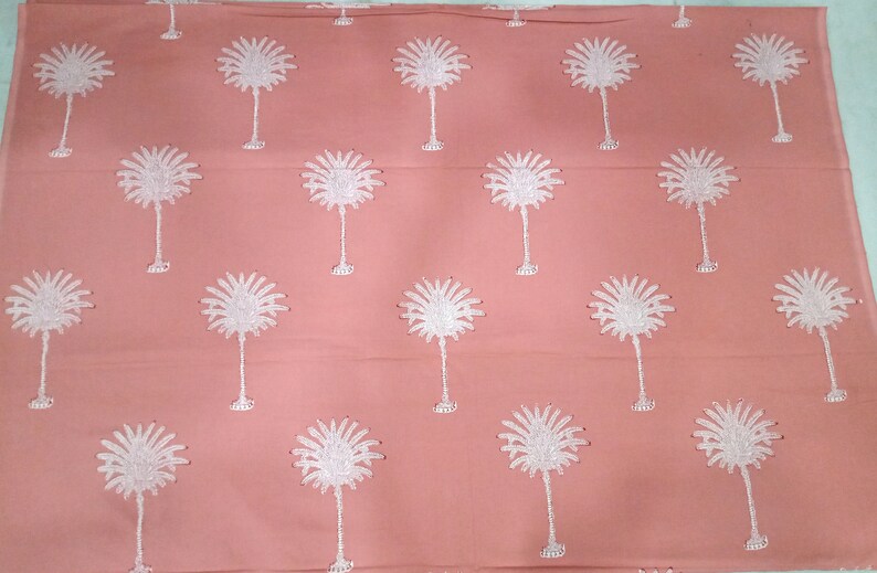 May include: A coral-colored fabric with a repeating pattern of white palm tree designs. The palm trees are embroidered with detailed fronds and slender trunks, creating a tropical aesthetic. The fabric is likely used for home decor or apparel.