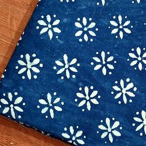 Pode incluir: Uma pe&ccedil;a de tecido azul-escuro com um padr&atilde;o repetitivo de formas florais brancas de seis p&eacute;talas e pequenos pontos brancos dispostos em grelha. O tecido tem um aspeto ligeiramente manchado e tingido &agrave; m&atilde;o com um design geom&eacute;trico.