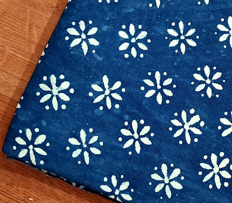 Pode incluir: Uma pe&ccedil;a de tecido azul-escuro com um padr&atilde;o repetitivo de formas florais brancas de seis p&eacute;talas e pequenos pontos brancos dispostos em grelha. O tecido tem um aspeto ligeiramente manchado e tingido &agrave; m&atilde;o com um design geom&eacute;trico.