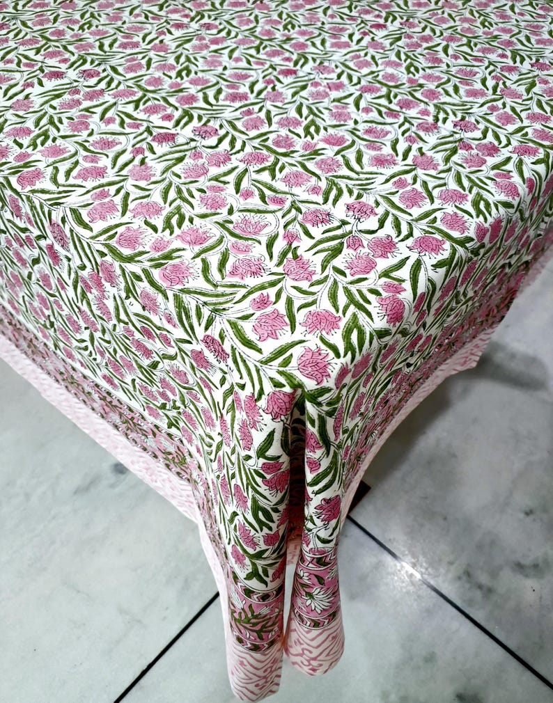May include: A white tablecloth featuring a repeating pattern of pink flowers and green leaves. The tablecloth has a pink border with a geometric design. The floral pattern covers the entire surface of the tablecloth.