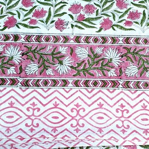 May include: A folded textile with a floral pattern in pink, green, and white. The design includes rows of flowers, geometric shapes, and decorative borders. The fabric is likely cotton or linen.