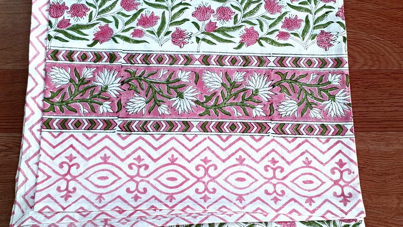 May include: A folded textile with a floral pattern in pink, green, and white. The design includes rows of flowers, geometric shapes, and decorative borders. The fabric is likely cotton or linen.