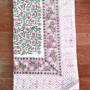 May include: A white and pink floral patterned textile. The design features pink flowers and green leaves on a white background. Borders include pink and white geometric patterns. The textile is likely a tablecloth or fabric.