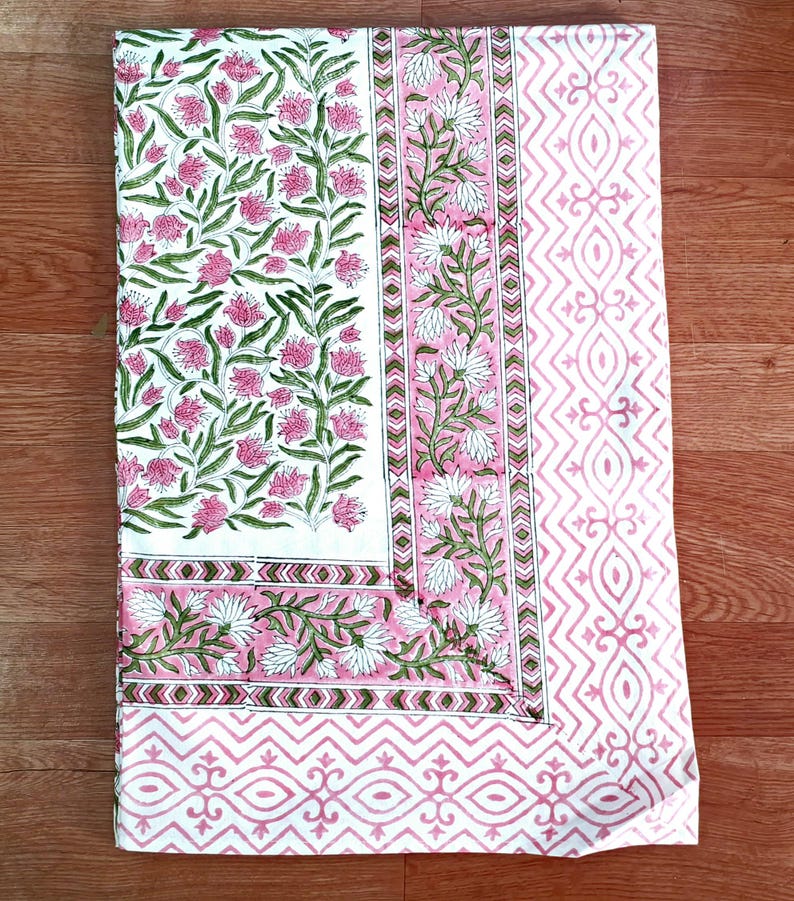 May include: A white and pink floral patterned textile. The design features pink flowers and green leaves on a white background. Borders include pink and white geometric patterns. The textile is likely a tablecloth or fabric.