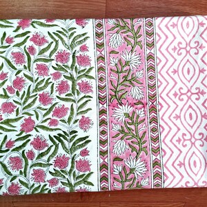 May include: A folded textile with a floral pattern. The design features pink flowers and green leaves on a white background. A pink and white geometric pattern is on the right side, and a pink and white floral stripe is in the middle.