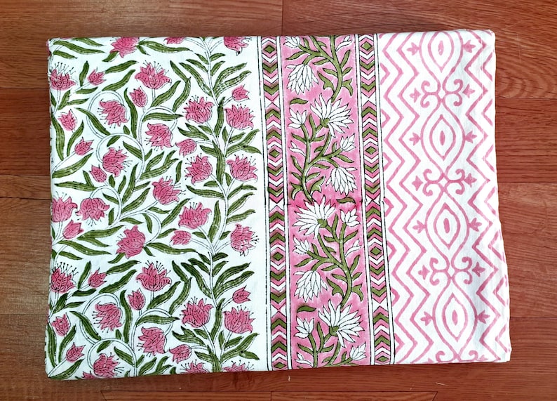 May include: A folded textile with a floral pattern. The design features pink flowers and green leaves on a white background. A pink and white geometric pattern is on the right side, and a pink and white floral stripe is in the middle.