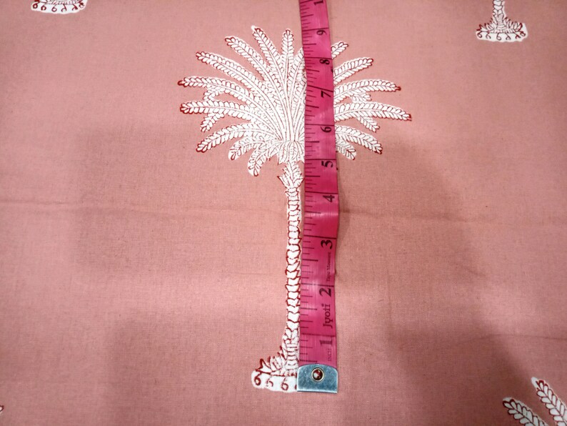 May include: Pink fabric with a repeating white and red palm tree pattern. A pink measuring tape shows a measurement of approximately 5 inches. The fabric is likely used for sewing or crafting projects.