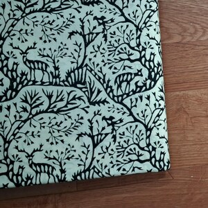 May include: A piece of fabric featuring a repeating pattern of black silhouettes of deer and stylized trees printed on a light mint green background. The design showcases multiple deer figures interspersed among intricate, branching tree motifs.