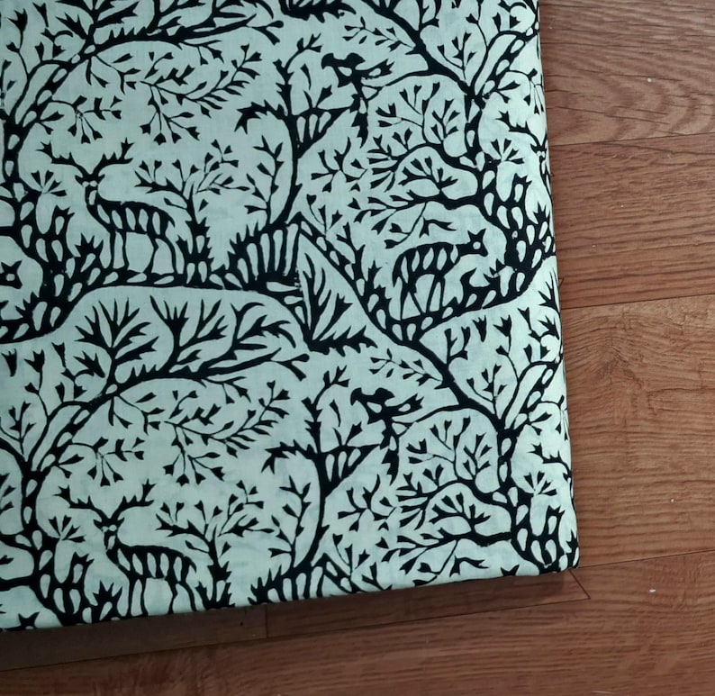 May include: A piece of fabric featuring a repeating pattern of black silhouettes of deer and stylized trees printed on a light mint green background. The design showcases multiple deer figures interspersed among intricate, branching tree motifs.