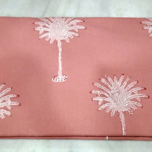 May include: A folded coral-colored fabric featuring a repeating pattern of white embroidered palm trees. The palm trees have detailed fronds and trunks, creating a tropical design. The fabric is likely cotton or linen.
