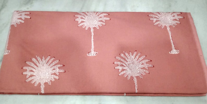 May include: A folded coral-colored fabric featuring a repeating pattern of white embroidered palm trees. The palm trees have detailed fronds and trunks, creating a tropical design. The fabric is likely cotton or linen.