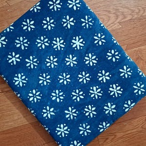 Pode incluir: Uma pe&ccedil;a de tecido dobrada com um fundo azul profundo e um padr&atilde;o repetitivo de flores brancas de seis p&eacute;talas. O tecido apresenta uma estampa floral consistente e uniformemente espa&ccedil;ada por toda a superf&iacute;cie vis&iacute;vel do material.