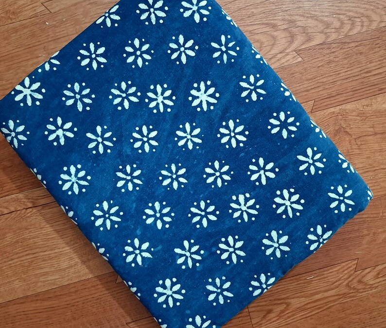 Pode incluir: Uma pe&ccedil;a de tecido dobrada com um fundo azul profundo e um padr&atilde;o repetitivo de flores brancas de seis p&eacute;talas. O tecido apresenta uma estampa floral consistente e uniformemente espa&ccedil;ada por toda a superf&iacute;cie vis&iacute;vel do material.