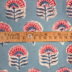 May include: Floral fabric with a repeating pattern of pink and blue stylized flowers on a light blue background. A wooden ruler shows measurements in inches. The fabric is likely cotton or a similar material.