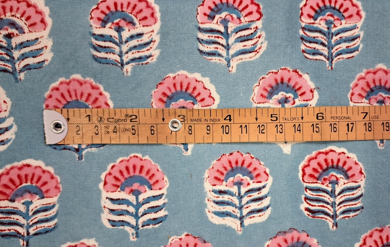 May include: Floral fabric with a repeating pattern of pink and blue stylized flowers on a light blue background. A wooden ruler shows measurements in inches. The fabric is likely cotton or a similar material.