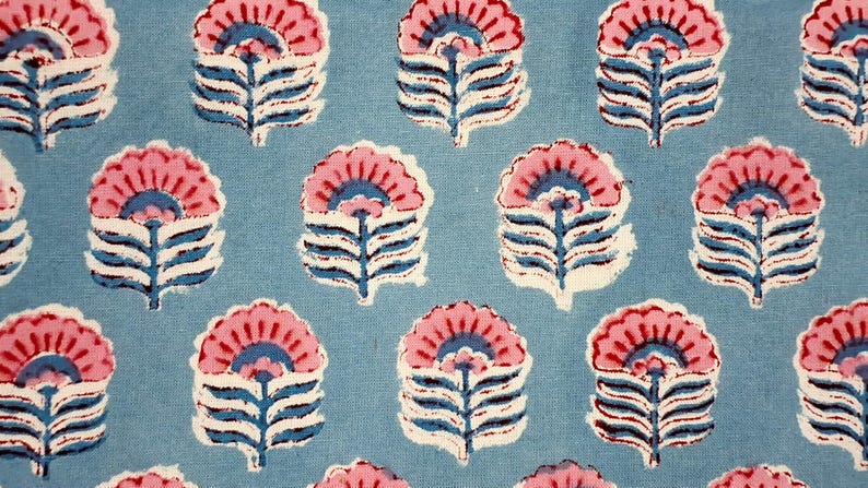 May include: A close-up of a fabric featuring a repeating floral design. The flowers have pink petals, blue centres, and white and blue stems, set against a light blue background. The design is reminiscent of block printing.
