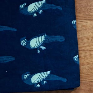 May include: A dark navy blue fabric featuring a repeating pattern of stylised birds in a lighter blue shade. Each bird is depicted in profile with a prominent wing, a long tail, and a small crest on its head, arranged in a consistent horizontal layout.