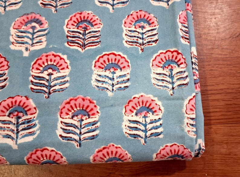 May include: Close-up of a folded fabric with a repeating floral pattern. The flowers are pink and white with blue accents, set against a light blue background. The fabric is likely cotton or linen.