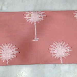 May include: A coral-colored fabric with a repeating pattern of white palm trees. The palm trees are embroidered with detailed fronds and trunks. The fabric is likely used for home decor or apparel.