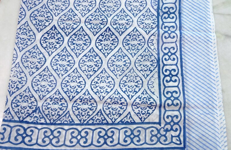 May include: A white cotton fabric with a blue and white geometric pattern. The fabric has a border with a repeating design.