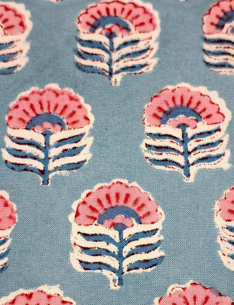 May include: Close-up of a light blue fabric with a repeating floral pattern. The flowers are pink with blue and white accents. The design has a block print style, creating a textured appearance. The fabric is likely cotton or linen.
