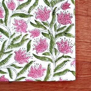 May include: A white fabric with a repeating floral pattern. The design features pink flowers with green leaves and stems. The fabric is placed on a wooden surface.