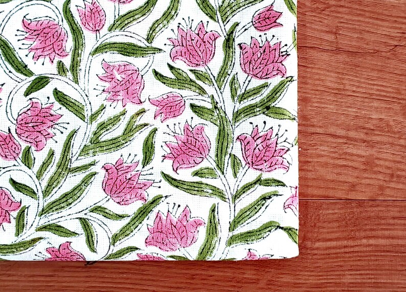 May include: A white fabric with a repeating floral pattern. The design features pink flowers with green leaves and stems. The fabric is placed on a wooden surface.
