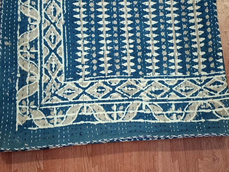 Puede incluir: Una colcha kantha azul y blanca con un patr&oacute;n geom&eacute;trico. La colcha est&aacute; cosida a mano y tiene un borde con flecos.