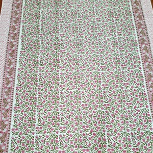 May include: A white tablecloth with a floral pattern in pink and green. The tablecloth has a border with a repeating geometric design in pink and brown. The floral pattern covers the majority of the tablecloth's surface.