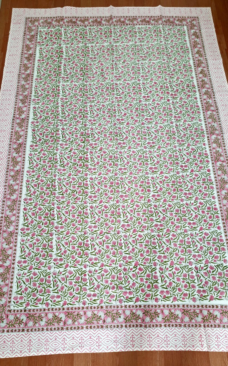May include: A white tablecloth with a floral pattern in pink and green. The tablecloth has a border with a repeating geometric design in pink and brown. The floral pattern covers the majority of the tablecloth's surface.
