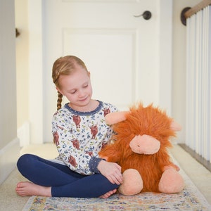 K&ouml;nnte beinhalten: Ein junges M&auml;dchen sitzt auf einem Teppich in einem Flur und h&auml;lt ein Pl&uuml;schtier in Form einer braunen und orangefarbenen Highland Cow. Sie tr&auml;gt ein lang&auml;rmliges Hemd in Wei&szlig; und Blau mit einem Muster aus Highland Cows und blaue Leggings.