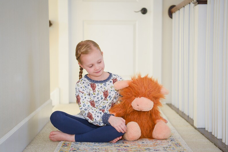 K&ouml;nnte beinhalten: Ein junges M&auml;dchen sitzt auf einem Teppich in einem Flur und h&auml;lt ein Pl&uuml;schtier in Form einer braunen und orangefarbenen Highland Cow. Sie tr&auml;gt ein lang&auml;rmliges Hemd in Wei&szlig; und Blau mit einem Muster aus Highland Cows und blaue Leggings.