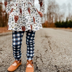 K&ouml;nnte beinhalten: Ein junges M&auml;dchen tr&auml;gt ein wei&szlig;es Kleid mit braunem Hochlandrind-Druck und blau-wei&szlig; karierten Leggings. Sie tr&auml;gt braune Ballerinas mit einem Riemen &uuml;ber dem Oberteil.