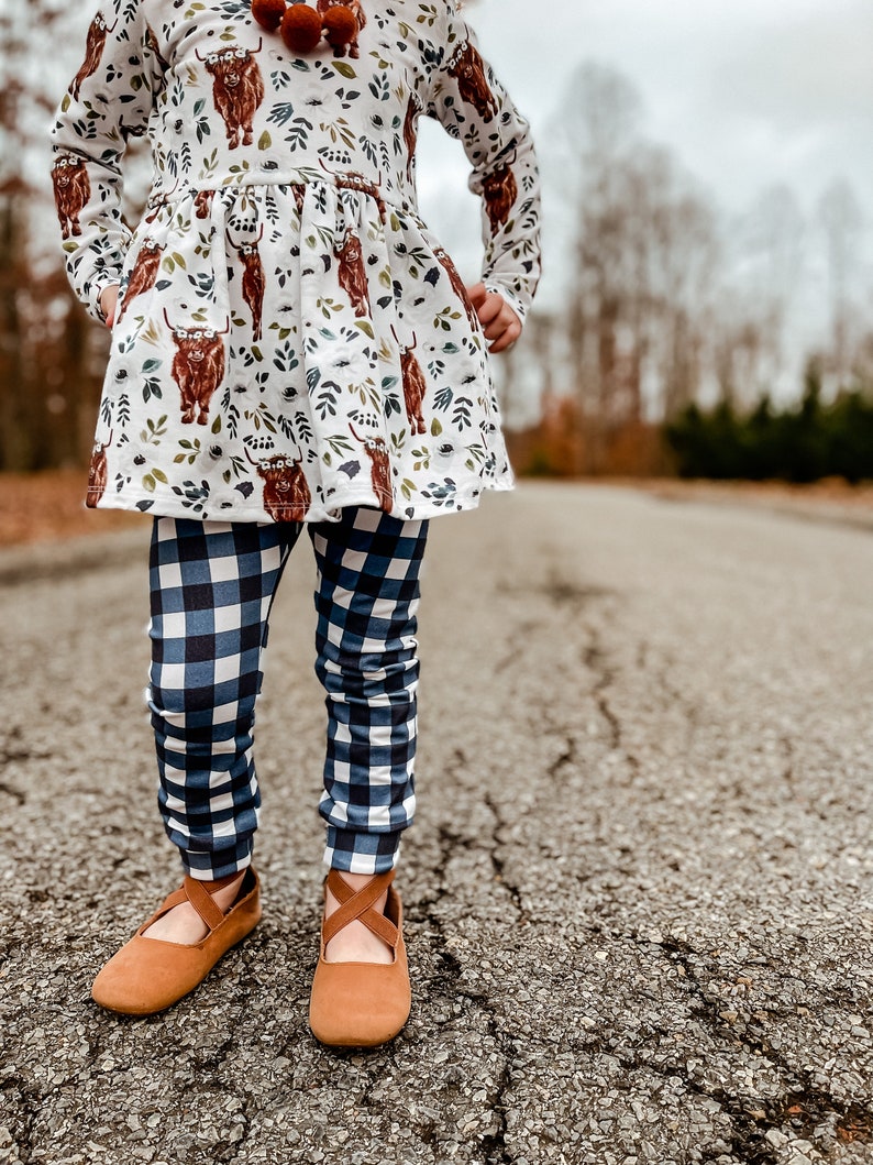 K&ouml;nnte beinhalten: Ein junges M&auml;dchen tr&auml;gt ein wei&szlig;es Kleid mit braunem Hochlandrind-Druck und blau-wei&szlig; karierten Leggings. Sie tr&auml;gt braune Ballerinas mit einem Riemen &uuml;ber dem Oberteil.