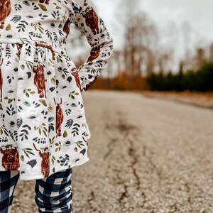 K&ouml;nnte beinhalten: Ein junges M&auml;dchen tr&auml;gt ein wei&szlig;es Kleid mit braunem Hochlandrind-Druck und blau-wei&szlig; karierten Leggings. Das Kleid hat lange &Auml;rmel und einen R&uuml;schen-Saum. Das M&auml;dchen steht auf einer rissigen Asphaltstra&szlig;e.