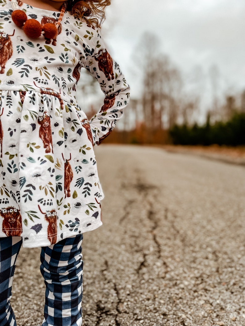 K&ouml;nnte beinhalten: Ein junges M&auml;dchen tr&auml;gt ein wei&szlig;es Kleid mit braunem Hochlandrind-Druck und blau-wei&szlig; karierten Leggings. Das Kleid hat lange &Auml;rmel und einen R&uuml;schen-Saum. Das M&auml;dchen steht auf einer rissigen Asphaltstra&szlig;e.