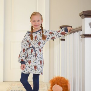 K&ouml;nnte beinhalten: Ein junges M&auml;dchen mit langen Z&ouml;pfen tr&auml;gt ein wei&szlig;es Kleid mit einem braunen und blauen Hochlandrind-Druck, dunkelblaue Leggings und steht auf einem gemusterten Teppich. Sie h&auml;lt sich mit einer Hand an einem wei&szlig;en Gel&auml;nder fest und ein gro&szlig;es, orangefarbenes, flauschiges Stofftier mit einer rosa Nase sitzt auf dem Teppich neben ihr.