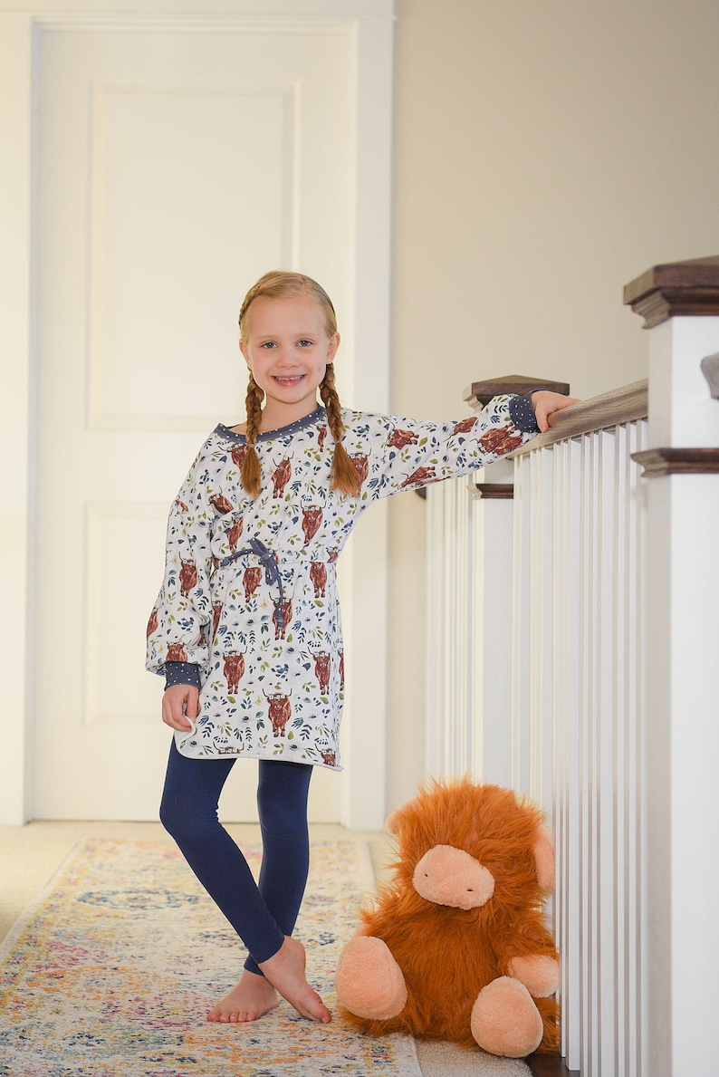K&ouml;nnte beinhalten: Ein junges M&auml;dchen mit langen Z&ouml;pfen tr&auml;gt ein wei&szlig;es Kleid mit einem braunen und blauen Hochlandrind-Druck, dunkelblaue Leggings und steht auf einem gemusterten Teppich. Sie h&auml;lt sich mit einer Hand an einem wei&szlig;en Gel&auml;nder fest und ein gro&szlig;es, orangefarbenes, flauschiges Stofftier mit einer rosa Nase sitzt auf dem Teppich neben ihr.