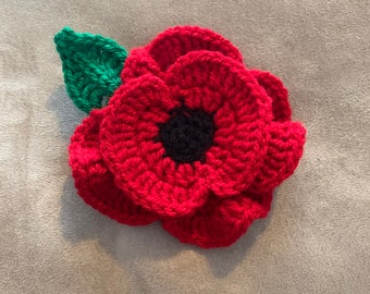 May include: A handcrafted red crochet poppy with a black centre and a green leaf. The poppy is a symbol of remembrance. The flower is made of yarn and has a textured appearance.