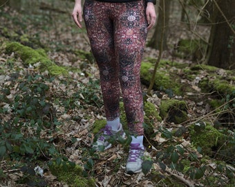 May include: A woman wearing a black top and patterned leggings stands in a woodland setting. She has a hair tie and glasses on.