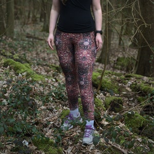May include: A woman wearing a black shirt and patterned leggings stands in a forest. She is wearing a hair tie and glasses.