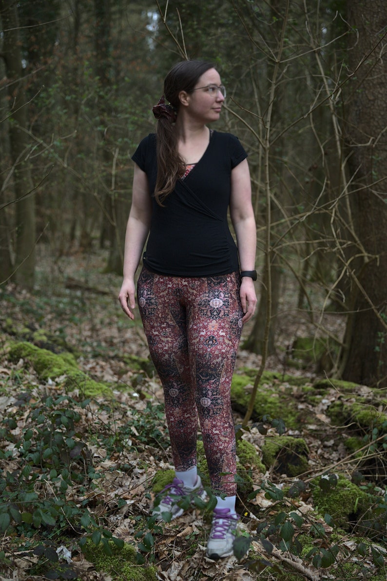 May include: A woman wearing a black shirt and patterned leggings stands in a forest. She is wearing a hair tie and glasses.