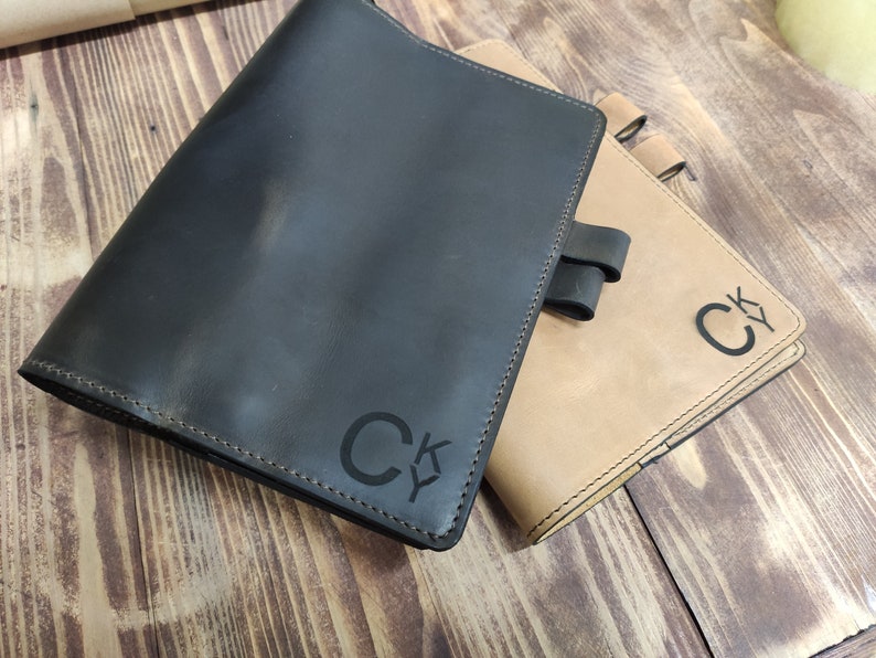 Puede incluir: Dos diarios encuadernados en cuero con las iniciales personalizadas "CK" grabadas en las tapas. El diario de la izquierda es negro y el diario de la derecha es marr&oacute;n.