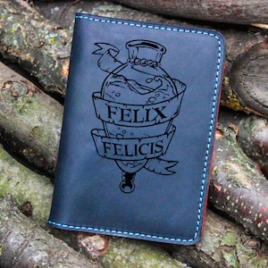 Könnte beinhalten: Ein blauer Leder-Reisepasshalter mit einem genähten Rand. Der Halter zeigt ein eingraviertes Design einer Flasche mit dem Text "Felix Felicis" auf einem Band.