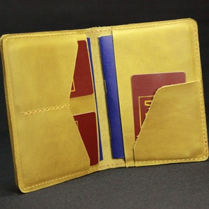 May include: A yellow leather passport holder with two pockets for passports or other documents. The holder is open and shows a blue passport and two red cards inside.